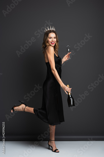 full length of happy woman in black slip dress and tiara with diamonds holding bottle of champagne and glass on grey