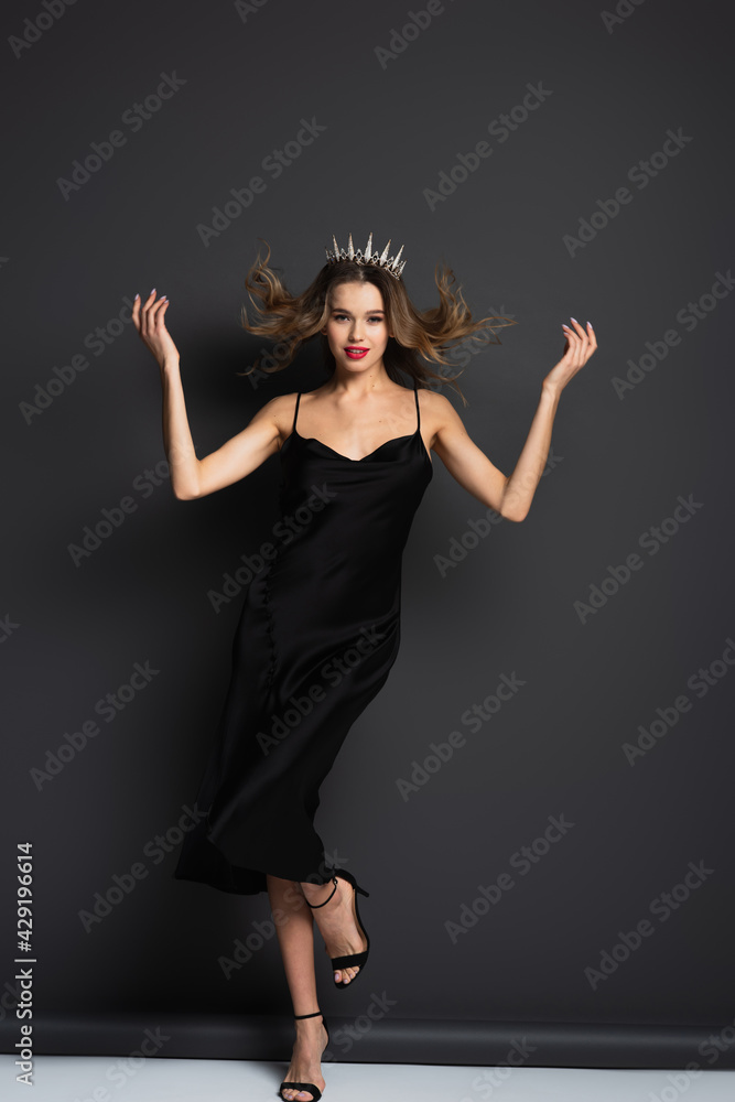 full length of elegant woman in black slip dress and tiara on grey