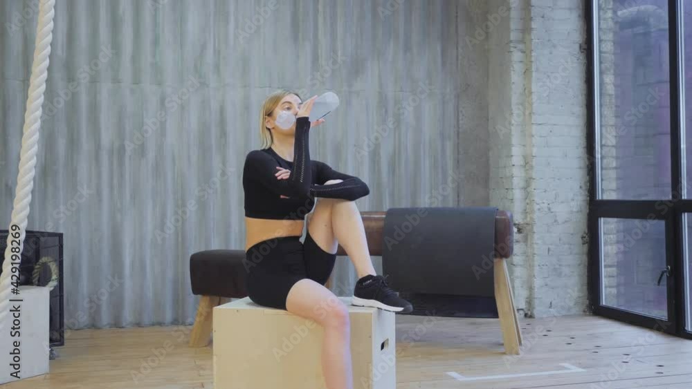 A Young Blonde Athletic Woman Drinks Water From A Bottle After An Exercise, Sits on a Jump Box, And Is Dressed in a Black Top and Leggings in a Light Gym A Young Blonde Athletic Woman Drinks Water From A Bottle After An Exercise, Sits on a Jump Box, And Is Dressed in a Black Top and Leggings in a Light Gym