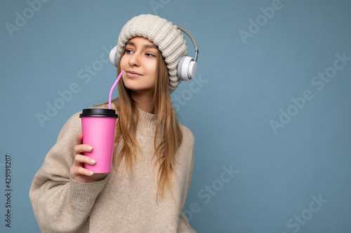 Photo shot of Beautiful young happy blonde woman wearing knitted hat beige sweater and white wireless headsets listening to music isolated over blue background holding paper tes cup for mockup