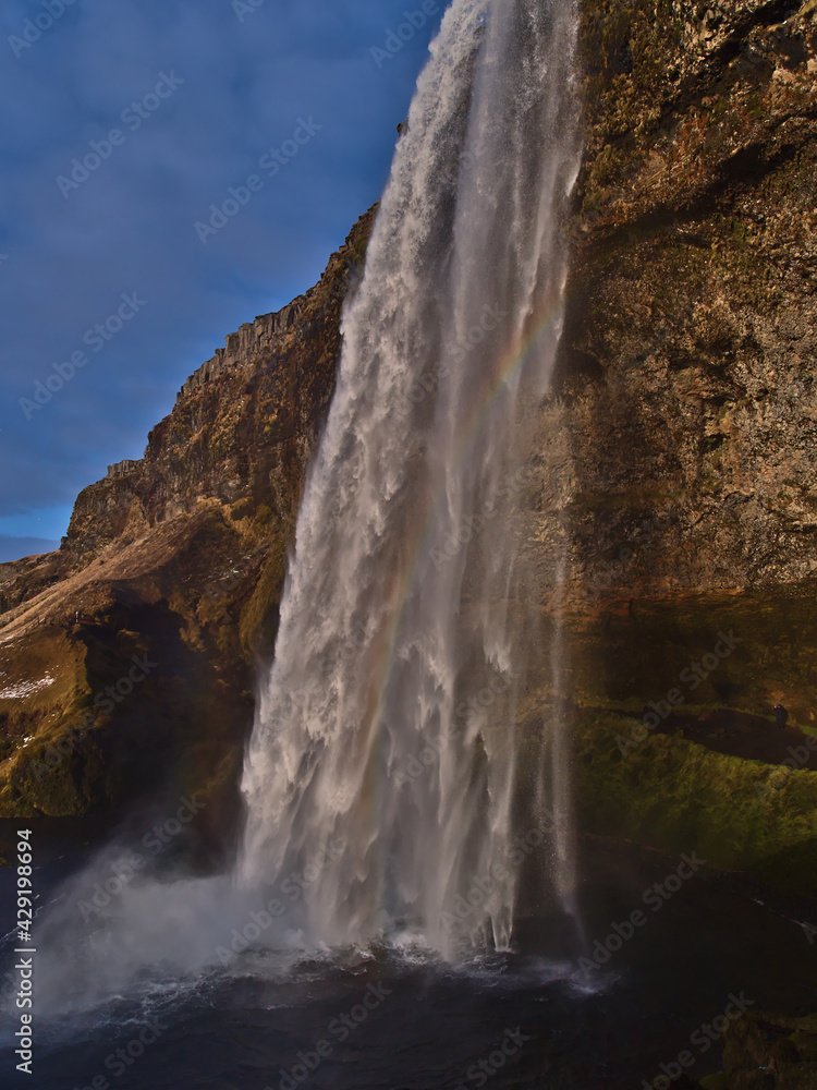 Beautiful side view of famous waterfall Seljalandsfoss on the southern coast of Iceland near ring road with colorful rainbow in between in the evening sun in winter season.