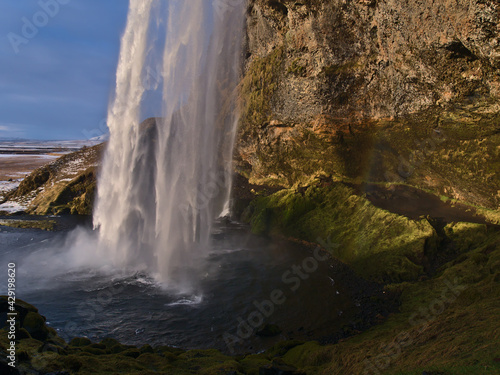 Stunning view of the bottom of famous waterfall Seljalandsfoss in the south of Iceland with moss-covered colorful rock face in the beautiful evening light in winter season.