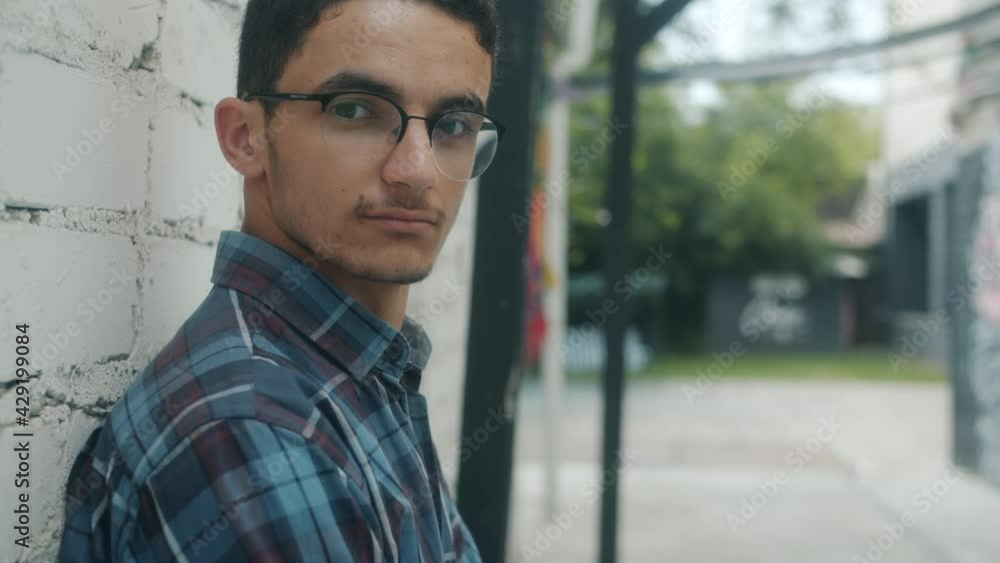 Serious Middle Eastern man is turning to camera standing near brick wall outdoors on summer day wearing casual clothes and glasses. People and background concept. Serious Middle Eastern man is turning to camera standing near brick wall outdoors on summer day wearing casual clothes and glasses. People and background concept.