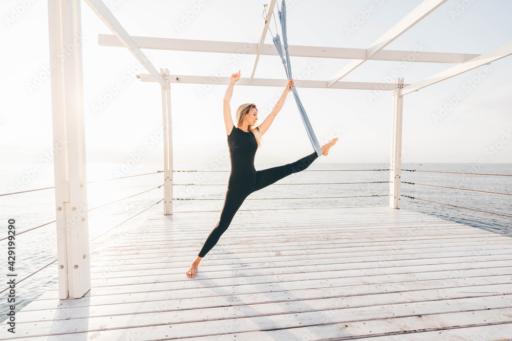 Relaxed young woman with blonde loose hair in sportswear does stretching exercises with hanging blue sling on empty pier near tranquil sea water at sunset