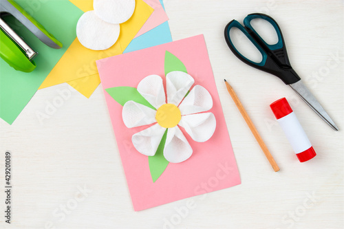 A flower made of cotton pads. Colored paper, stapler, scissors, scissors, pencil on a white table. Children's craft card for mother's day. Step-by-step instruction. Step 12.