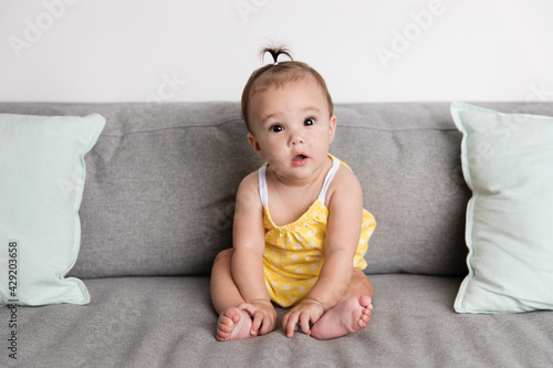 Cute baby girl sitting on sofa looking at camera with suprised expression photo