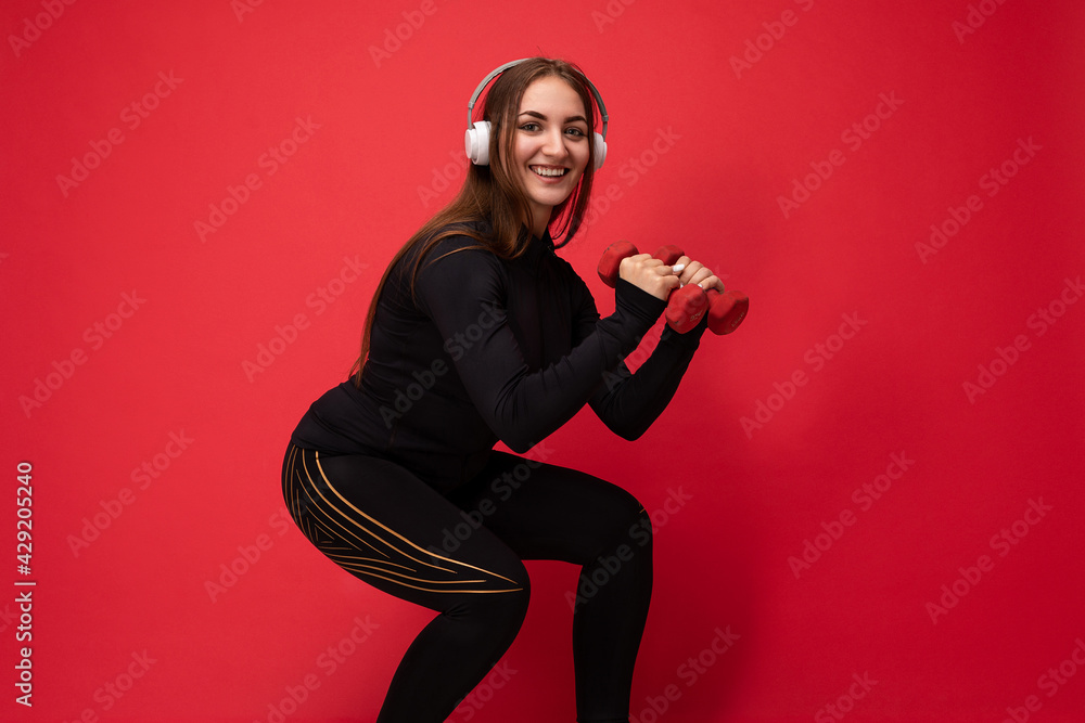 Side-profile photo of beautiful positive smiling young brunette woman wearing black sport clothes isolated on red background wall squating using dumbbells wearing white bluetooth earphones listening