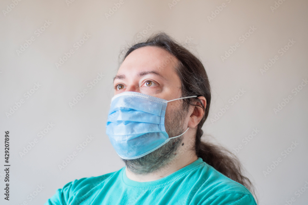 An adult male wearing a protective medical mask looks to the upper left upper corner. Close-up of a man against a light background. An adult male wearing a protective medical mask looks to the upper left upper corner. Close-up of a man against a light background.