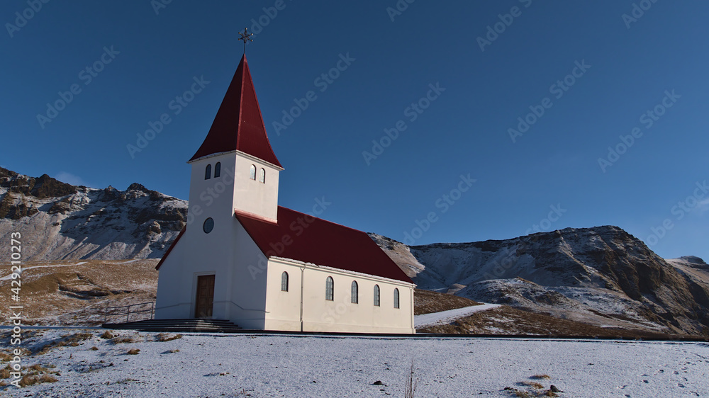 Beautiful view of the small church of village Vík í Mýrdal located on the southern coast of Iceland with white wall and red colored roof on sunny winter day with clear sky and rugged mountains.