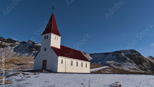 Beautiful view of the small church of village Vík í Mýrdal located on the southern coast of Iceland with white wall and red colored roof on sunny winter day with clear sky and rugged mountains.