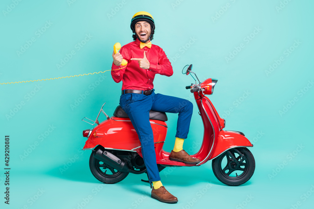 Full size photo of young funny smiling man riding moped point finger cord phone recommend isolated on turquoise color background Full size photo of young funny smiling man riding moped point finger cord phone recommend isolated on turquoise color background