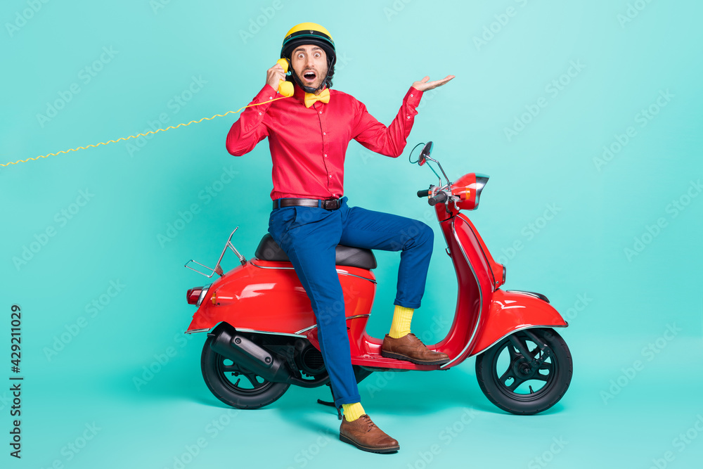 Full size photo of young shocked amazed man sit moped talking on telephone isolated on turquoise color background Full size photo of young shocked amazed man sit moped talking on telephone isolated on turquoise color background