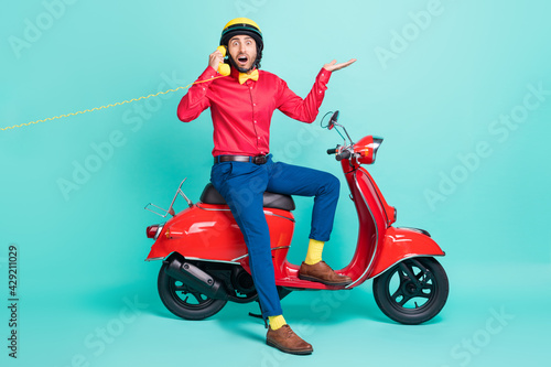 Full size photo of young shocked amazed man sit moped talking on telephone isolated on turquoise color background