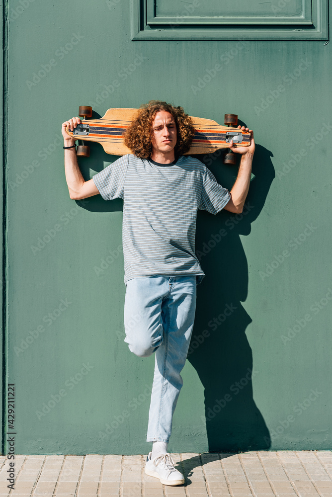 attractive caucasian freckled young man holding skateboard on shoulders. He is resting on a green wall.