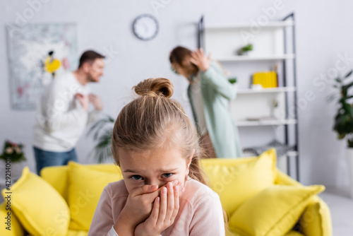 Scared child standing near parents quarrelling in living room on blurred background.