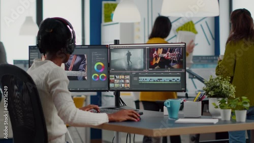 Black videographer putting on headset editing movie using post production software working in creative start up agency office. African editor processing video footage on computer with dual displays