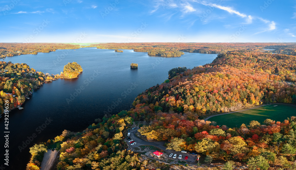 Aerial view of colorful fall forest by the fresh water lake - bright yellow, red, orange, green  trees. Blue sky, sunny day. Lion's Lookout, Muskoka, Northern Ontario, Canada.