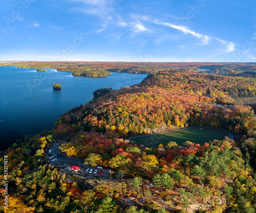 Aerial view of colorful fall forest by the fresh water lake - bright yellow, red, orange, green trees. Blue sky, sunny day. Lion's Lookout, Muskoka, Northern Ontario, Canada.