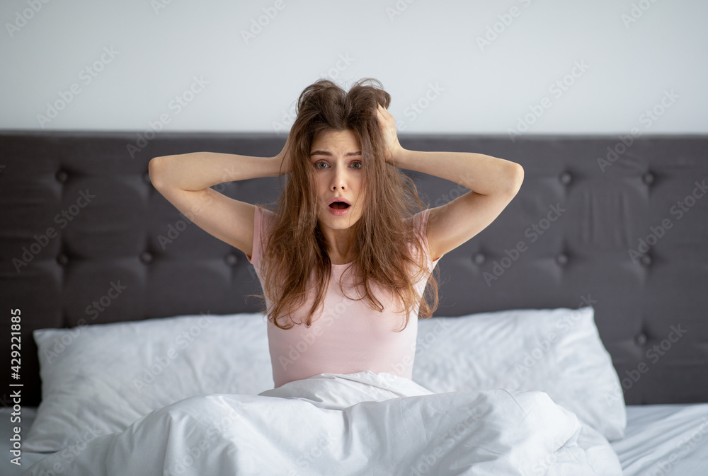 Shocked yound woman sitting in bed at home, grabbing her head in terror, oversleeping, being late to work Shocked yound woman sitting in bed at home, grabbing her head in terror, oversleeping, being late to work