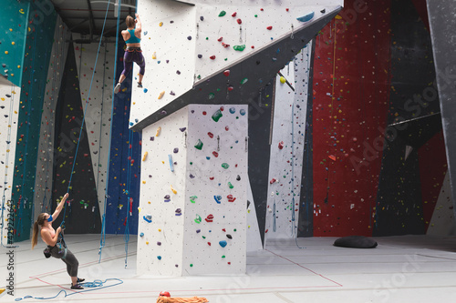 Two caucasian women wearing face masks using ropes to climb wall at indoor climbing gym photo