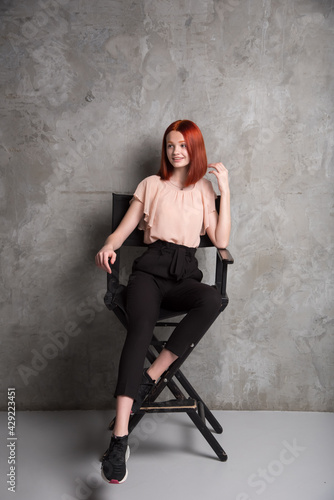 .The bright red-haired girl sits on the director's chair in full growth. Against the background of a concrete gray modern wall.