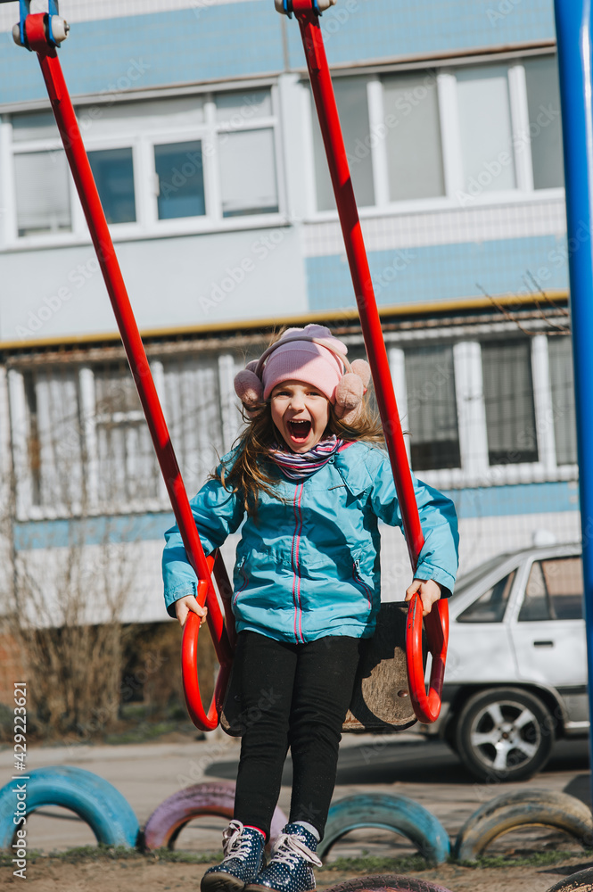 A funny, smiling girl, a preschool child rides, sitting on a metal red swing, on the playground. Happy childhood. A funny, smiling girl, a preschool child rides, sitting on a metal red swing, on the playground. Happy childhood.