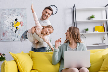 Smiling mother with laptop looking at family having fun at home.