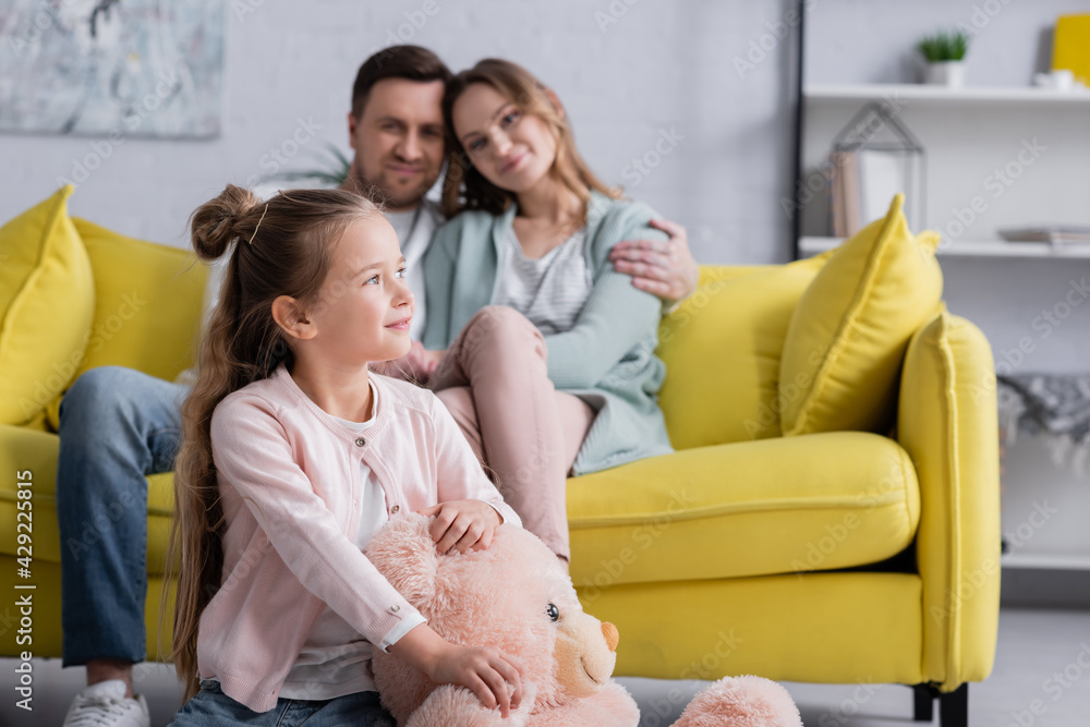 Positive kid holding soft toy near parents on blurred background. Positive kid holding soft toy near parents on blurred background.
