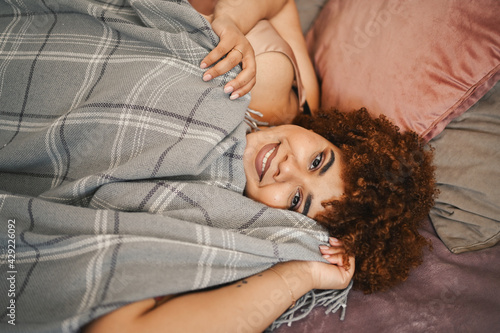 Beautiful curvy plus size African black woman afro hair lying on bed with grey plaid blanket cozy bedroom interior design. Body imperfection  body acceptance  body positive and diversity concept.