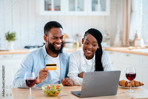 Food Ordering. Happy Black Spouses Using Laptop And Card In Kitchen