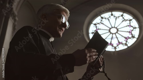 Low angle view of old Caucasian pastor wearing black robe and white collar standing in Lutheran church with Holy Bible and rosary beads in hands and praying