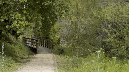 A dirt walk with a brown wooden bridge and green vegetation around it with a stone house on one side and trees. hiking trail through the nature center and abandoned stone house. route in ponteareas