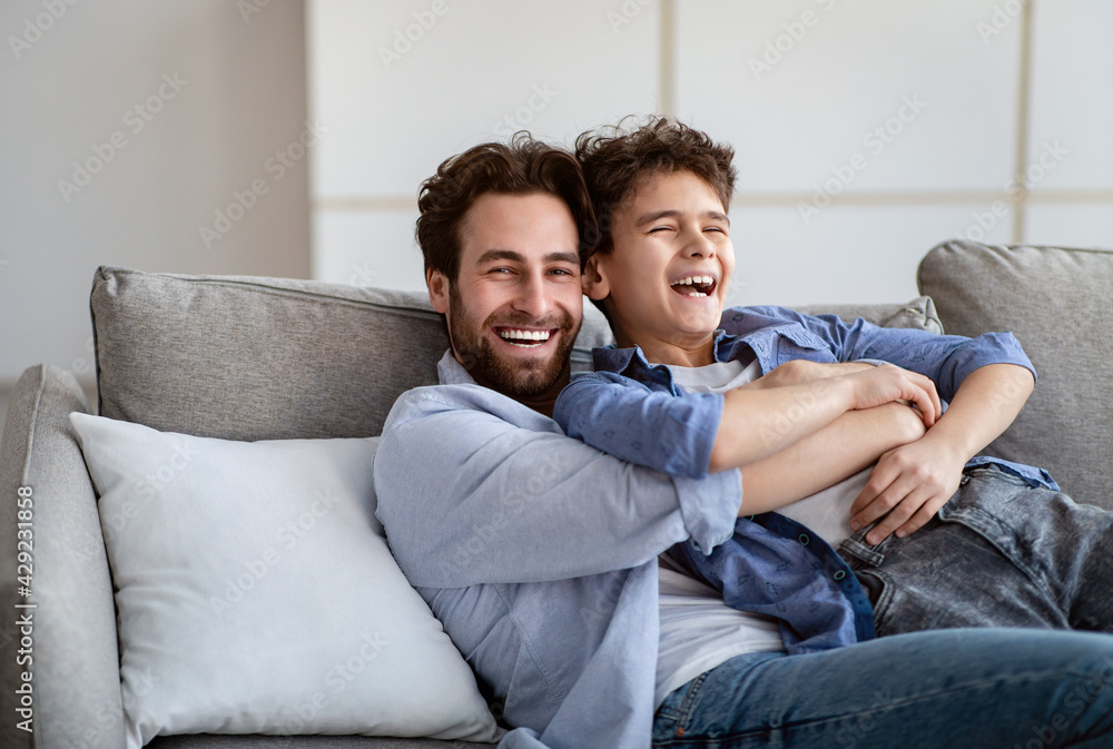 Fatherhood concept. Cheerful dad embracing son, tickling and laughing while resting on couch at home in living room Fatherhood concept. Cheerful dad embracing son, tickling and laughing while resting on couch at home in living room