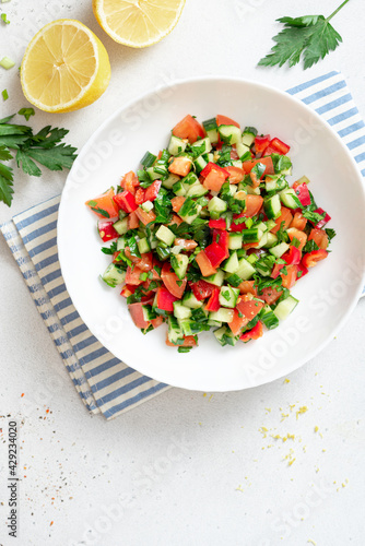 Vegetable salad on a white plate on a light background. Salad with tomatoes, cucumbers, peppers, parsley and lemon. Vegan or diet food. Copy space for text.