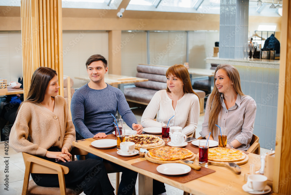 A group of young cheerful friends is sitting in a cafe talking and eating pizza. Lunch at the pizzeria. A group of young cheerful friends is sitting in a cafe talking and eating pizza. Lunch at the pizzeria.