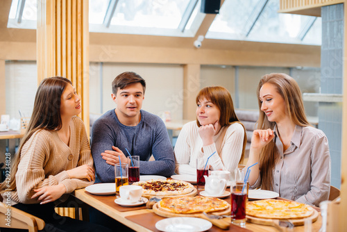 A group of young cheerful friends is sitting in a cafe talking and eating pizza. Lunch at the pizzeria.