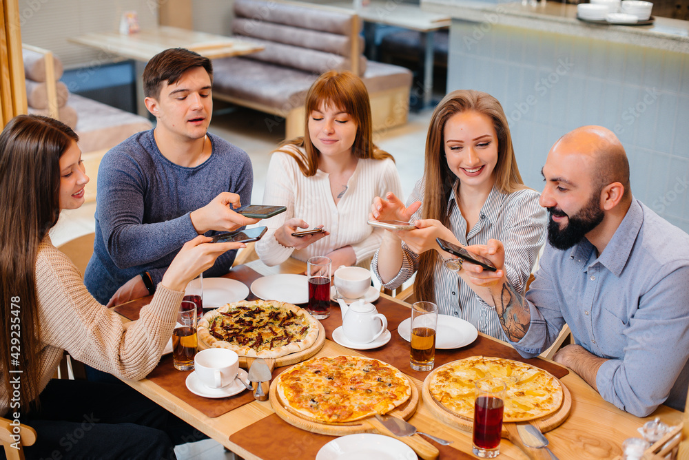 A group of young cheerful friends is sitting in a cafe talking and taking selfies on the phone. Lunch at the pizzeria.