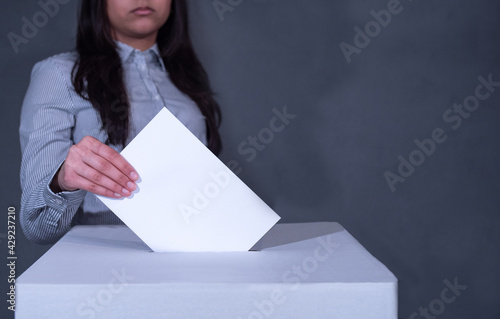 Junge Frau im gestreiften Hemd steht hinter einer Wahlbox und wirft ihren Stimmzettel hinein. photo