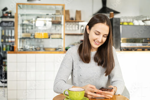 Smiling young brunette woman enjoys a morning coffee sitting in the cozy cafe and chatting on the smartphone  female student has a break with phone and hot drink in the cafeteria