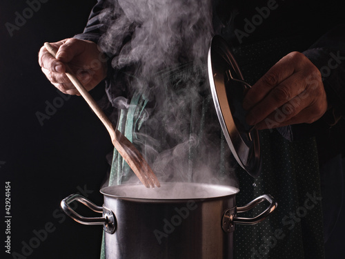 The cook is in the kitchen. Close-up of an open saucepan lid and a chef's hand with a wooden fork on a dark green speckled apron background.