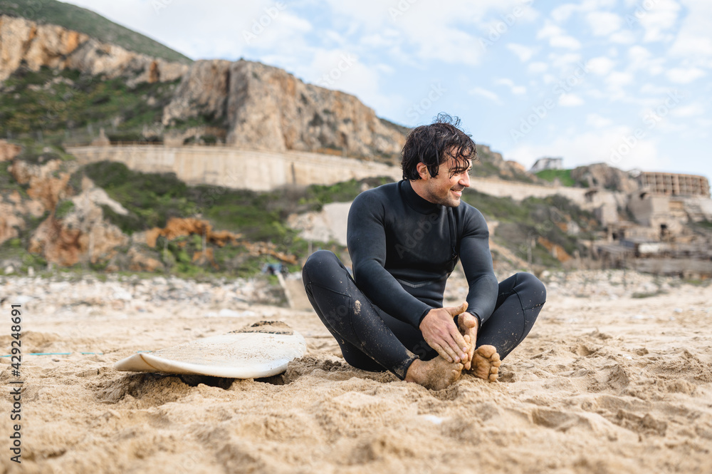 Young surfer man wearing diving suit sitting on the sand next to his surfboard resting after surfing.