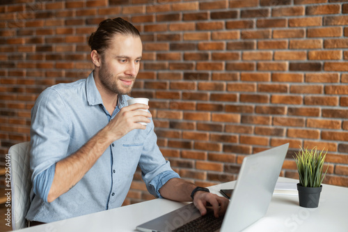 Side view a male freelancer drinking coffee, using a laptop for work in a loft style office. A millennial guy works on outsours, receiving emails, conducts correspondence with customer