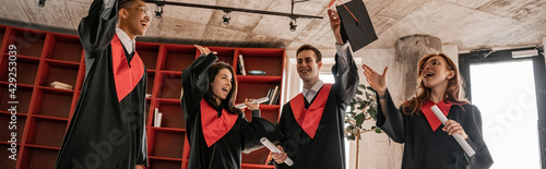 amazed interracial students in graduation gowns holding diploma, senior 2021, banner