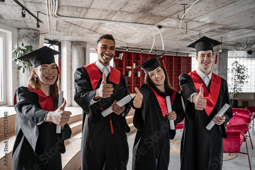 happy interracial students in graduation gowns and caps holding diploma and showing thumbs up, senior 2021