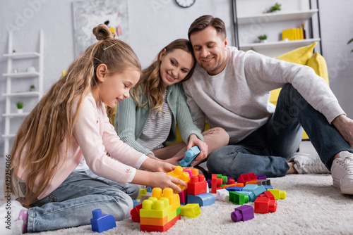 Smiling kid playing building blocks on carpet near blurred parents.