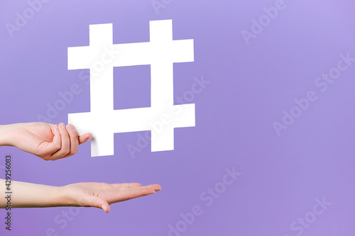 Concept of trendy social media posts and blogging. Female hands holding large big white hashtag sign, viral web content, internet promotion, isolated on purple studio background with copy space