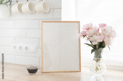 Vertical frame mockup on a wooden table in the kitchen. Glass vase with a bouquet of pink peonies and a cup of black coffee. Scandinavian style interior.