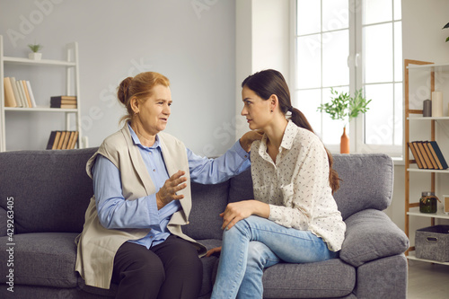 Supportive senior mother talking to her grown-up young daughter while sitting on sofa at home. Love, care, family support, mutual help and understanding concept