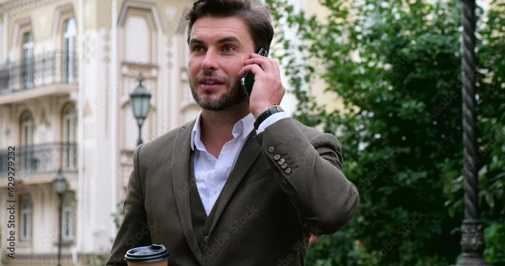 Attractive young freelancer talking by phone with Coffee.Handsome young businessman in stylish suit with paper cup of coffee using smartphone near business building. european street in the background Attractive young freelancer talking by phone with Coffee.Handsome young businessman in stylish suit with paper cup of coffee using smartphone near business building. european street in the background