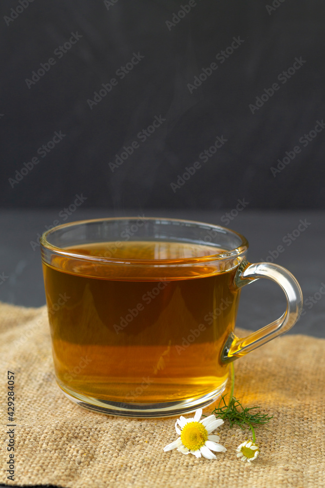 hot drinks. mug of chamomile tea on sackcloth. glass cup of iced Herbal chamomile tea on a black table with copy space. cup of tea with fresh flowers and green leaves on grey background. vertical hot drinks. mug of chamomile tea on sackcloth. glass cup of iced Herbal chamomile tea on a black table with copy space. cup of tea with fresh flowers and green leaves on grey background. vertical